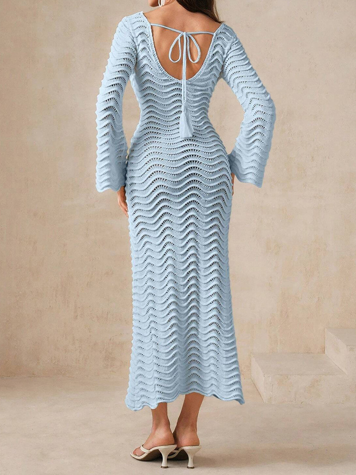 Back view of a woman wearing a light blue, long-sleeved, midi-length knit dress with a wave pattern, bell sleeves, and an open back with a tie closure.