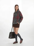 Full body shot of a woman in a plaid collared knit jacket, holding a dark leather tote bag.