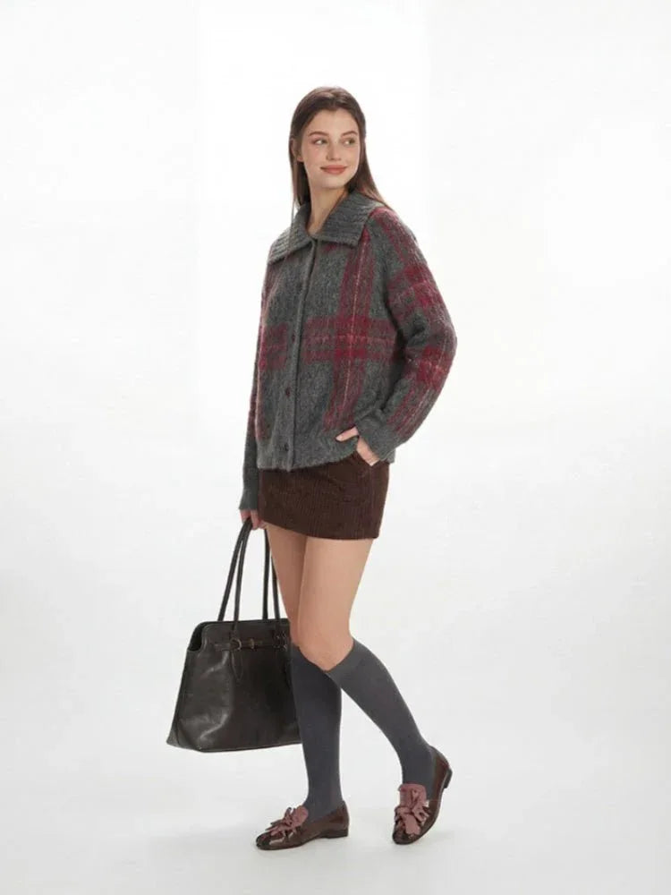 Full body shot of a woman in a plaid collared knit jacket, holding a dark leather tote bag.
