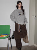 A full-body shot of a woman in a gray crew neck sweatshirt and brown pants.