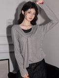 Woman posing with an arm raised, wearing a grey knit top with a draped neckline and layered effect.