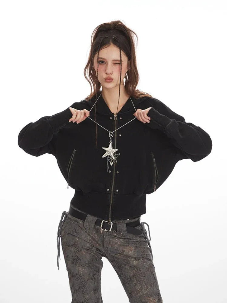 Woman posing with a black cropped hooded zip-up jacket and patterned pants, holding the neckline.