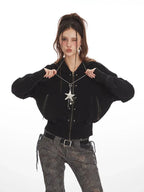 Woman posing with a black cropped hooded zip-up jacket and patterned pants, holding the neckline.