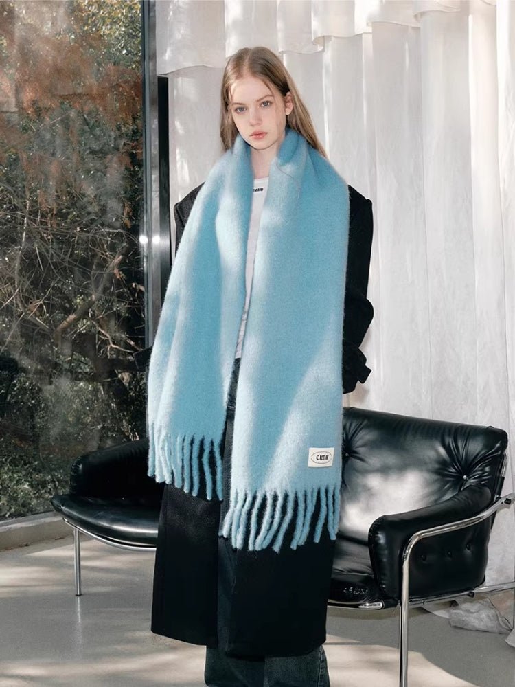 Woman wearing a light blue mohair blend scarf with fringe detailing and a white logo tag, draped over a black coat.