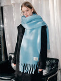 Woman wearing a light blue mohair blend scarf with fringe detailing and a white logo tag, draped over a black coat.
