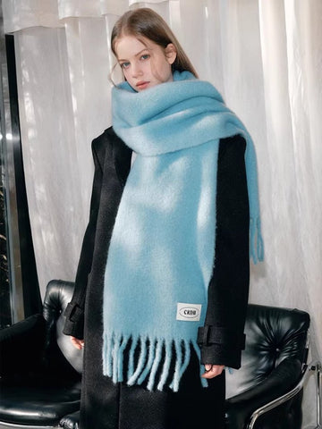 Woman wearing a light blue mohair blend scarf with fringe detailing and a white logo tag, draped over a black coat.