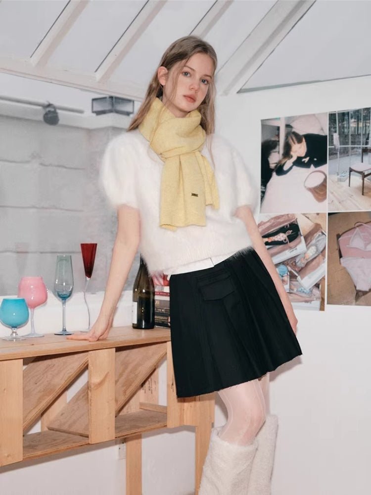 Woman with blonde hair wearing a white fuzzy short-sleeved sweater, a pale yellow scarf, and a black pleated skirt.