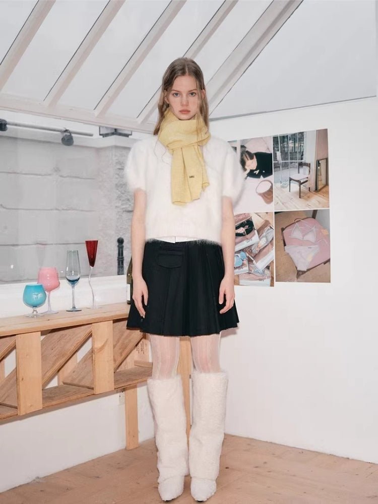 Full body shot of a model wearing a white fuzzy short-sleeved sweater, a pale yellow scarf, a black pleated skirt, and white fuzzy leg warmers.