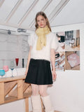 Model posing in a white fuzzy short-sleeved sweater, a pale yellow scarf, a black pleated skirt, and white fuzzy leg warmers in a minimalist room.