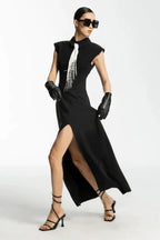 Woman in a black sleeveless collared maxi dress with a white tie and silver fringe detail, black leather gloves, and strappy heels, adjusting her dress.
