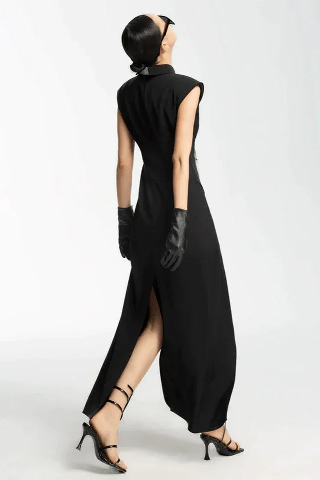 Rear view of a woman in a black sleeveless collared maxi dress with a white tie and silver fringe detail, black leather gloves, and strappy heels.