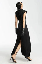 Rear view of a woman in a black sleeveless collared maxi dress with a white tie and silver fringe detail, black leather gloves, and strappy heels.