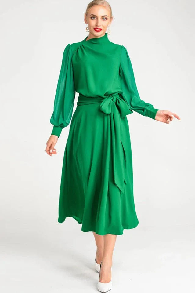 Woman wearing a green long-sleeved A-line dress with a mock neck and self-tie belt, angled front view