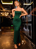 Woman in an emerald green satin mermaid dress with spaghetti straps and a square neckline, posing with a hand on her chest.
