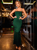 Woman in a floor-length emerald green satin mermaid dress with spaghetti straps and a square neckline, posing with hands on hips.