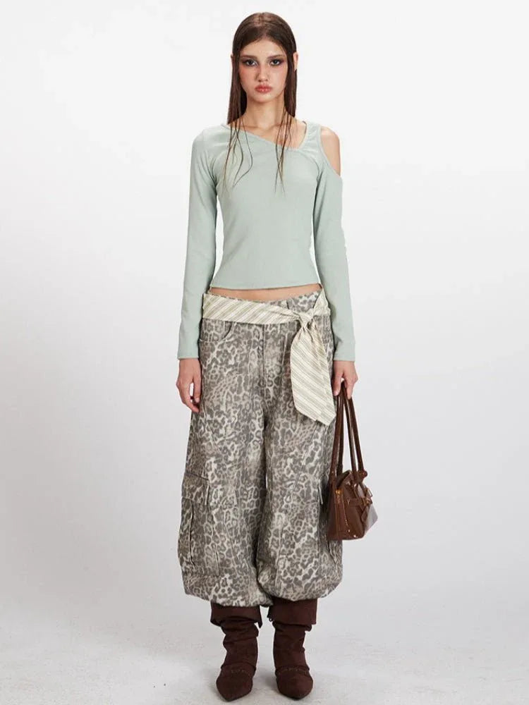 Woman wearing a light green long-sleeve ribbed knit top with asymmetrical neckline and cold shoulder cutouts, paired with leopard print wide-leg cargo pants and a striped sash.