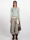Woman wearing a light green long-sleeve ribbed knit top with asymmetrical neckline and cold shoulder cutouts, paired with leopard print wide-leg cargo pants and a striped sash.