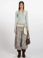 Woman wearing a light green long-sleeve ribbed knit top with asymmetrical neckline and cold shoulder cutouts, paired with leopard print wide-leg cargo pants and a striped sash.