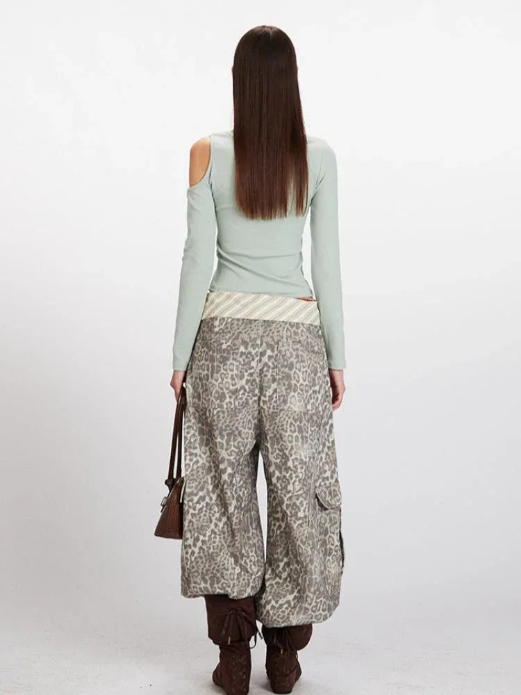 Woman wearing a light green long-sleeve ribbed knit top with asymmetrical neckline and cold shoulder cutouts, paired with leopard print wide-leg cargo pants and a striped sash.