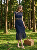 Woman in a navy blue lace midi dress with a high neckline and ruffled cap sleeves, standing in a forest with a picnic basket.