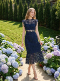 Woman in a navy blue lace midi dress with a high neckline and ruffled cap sleeves, smiling in a garden with hydrangeas.