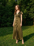 Woman in an olive green satin dress standing in a grassy area with trees