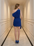 Back view of a woman in a royal blue sparkly one-shoulder mini dress with draped wrap skirt standing in a hallway.