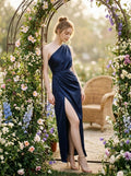 Woman in a navy blue satin one-shoulder gown with a thigh-high slit leaning against a floral archway.