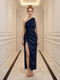 Woman in a navy blue satin one-shoulder gown with a thigh-high slit standing in a hallway.