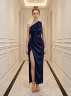 Woman in a navy blue satin one-shoulder gown with a thigh-high slit standing in a hallway.