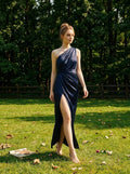 Woman in a navy blue satin one-shoulder gown with a thigh-high slit walking in a grassy field.