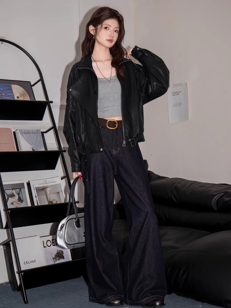 Full body shot of a woman in a black faux leather bomber jacket, grey crop top, and dark wash wide-leg jeans.