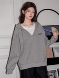 Woman in a grey hooded sweatshirt with an embroidered logo, styled with a white t-shirt underneath.