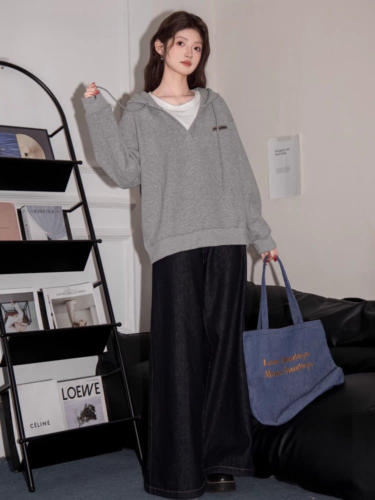 Full body shot of a woman in a grey hooded sweatshirt and dark wide-leg jeans, holding a blue tote bag.