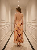 Rear view of a woman in a peach maxi dress with a tropical leaf print, showing an open back with crisscross straps.