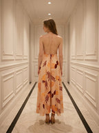 Rear view of a woman in a peach maxi dress with a tropical leaf print, showing an open back with crisscross straps.
