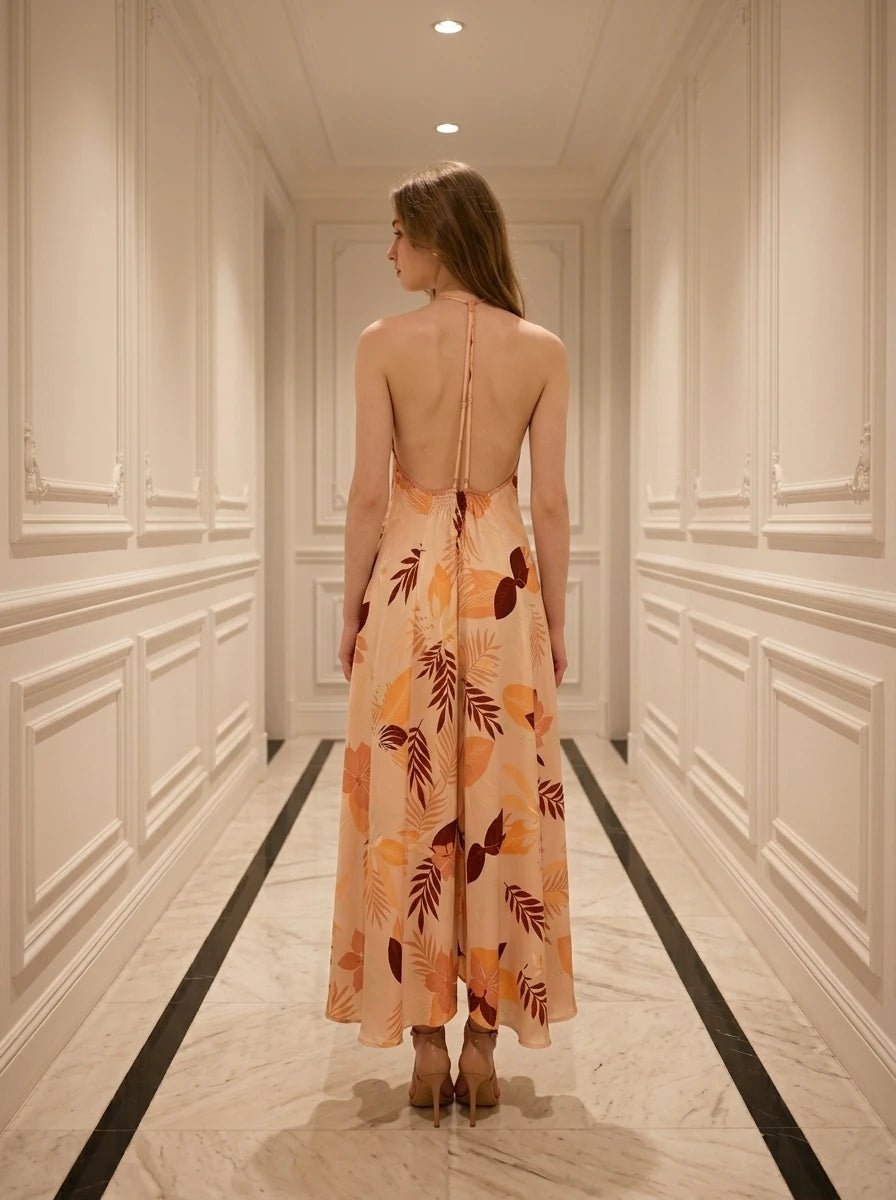 Rear view of a woman in a peach maxi dress with a tropical leaf print, showing an open back with crisscross straps.