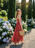 Woman wearing a red floral print maxi dress with spaghetti straps, holding a straw bag, walking in a garden with hydrangeas.