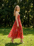Woman walking in a grassy area wearing a red floral print midi dress with pleats and cami straps.