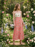 Woman wearing a coral midi dress with a patterned skirt and white bodice, standing in a garden with roses and hydrangeas.