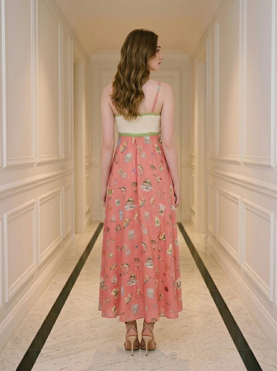 Woman wearing a coral midi dress with a white bodice featuring a bird and floral print, and a skirt with a repeating pattern of small illustrations of birds, cages, and buildings.