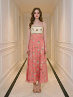 Woman wearing a coral midi dress with a white bodice featuring a bird and floral print, and a skirt with a repeating pattern of small illustrations of birds, cages, and buildings.