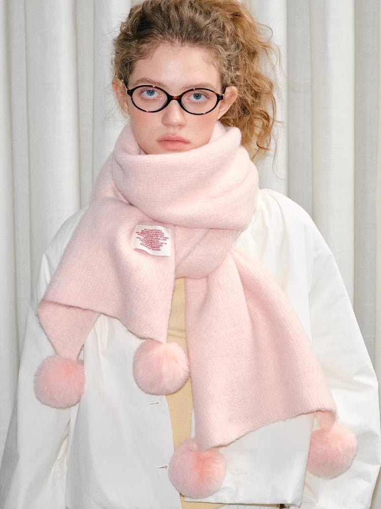 A woman wearing a pastel pink knitted scarf with fluffy pom-pom accents.