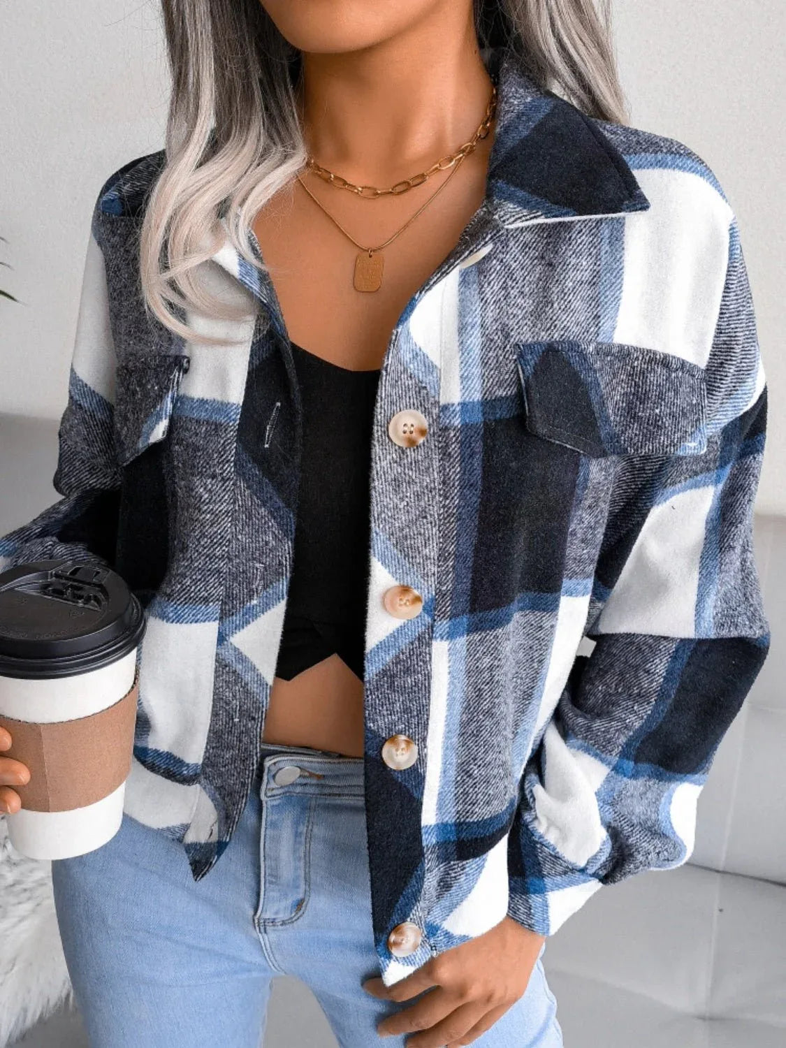 Woman holding a disposable coffee cup, wearing a plaid shirt jacket and jeans.