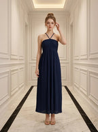 Woman in a navy blue pleated maxi dress with a halter neckline standing in a hallway.