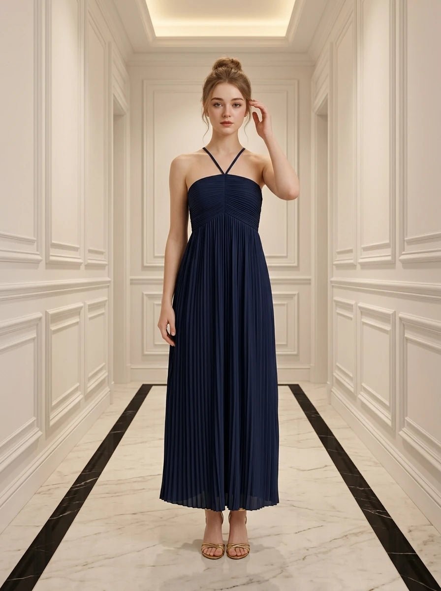 Woman in a navy blue pleated maxi dress with a halter neckline standing in a hallway.