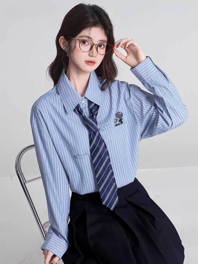 Full body shot of a woman in a light blue striped shirt, navy tie, and pleated skirt