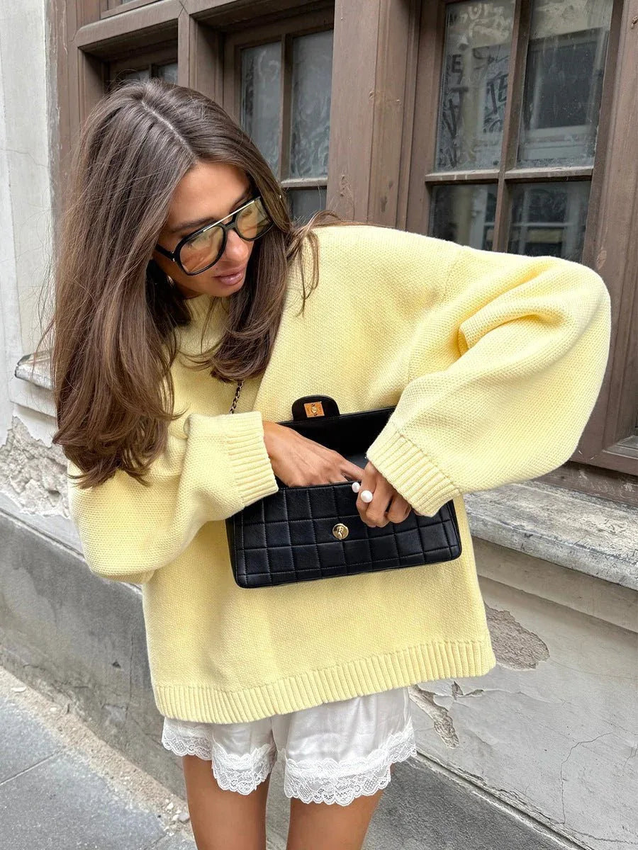 Woman wearing a light yellow crewneck sweater and white shorts, holding a black quilted clutch.