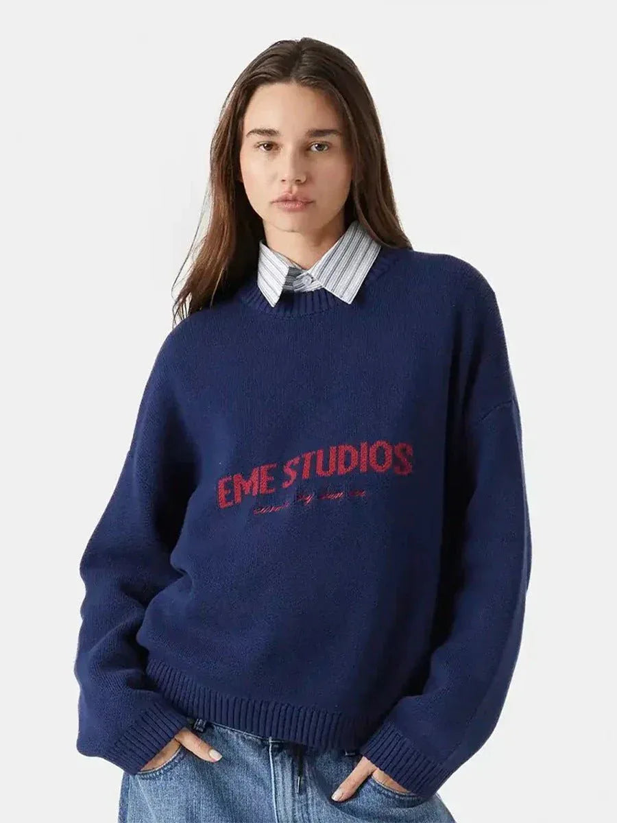 Woman wearing a navy blue crewneck sweater with red embroidered logo and a striped collared shirt underneath. Main image