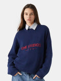 Woman wearing a navy blue crewneck sweater with red embroidered logo and a striped collared shirt underneath.